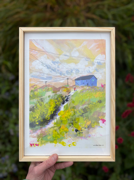 Framed painting of a landscape with a blue house held by a hand against a blurred natural background.
