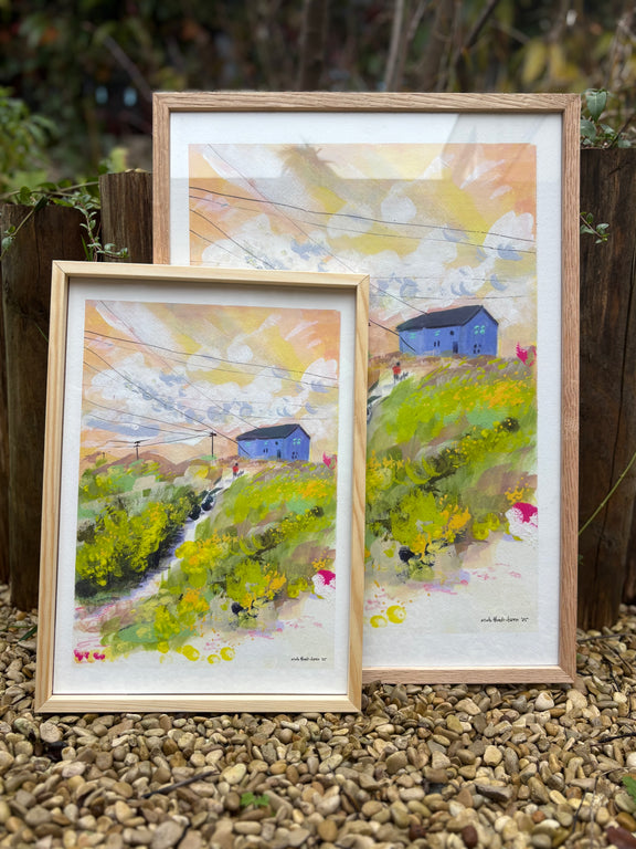 Two framed paintings of a landscape with a blue house on a pebble surface.