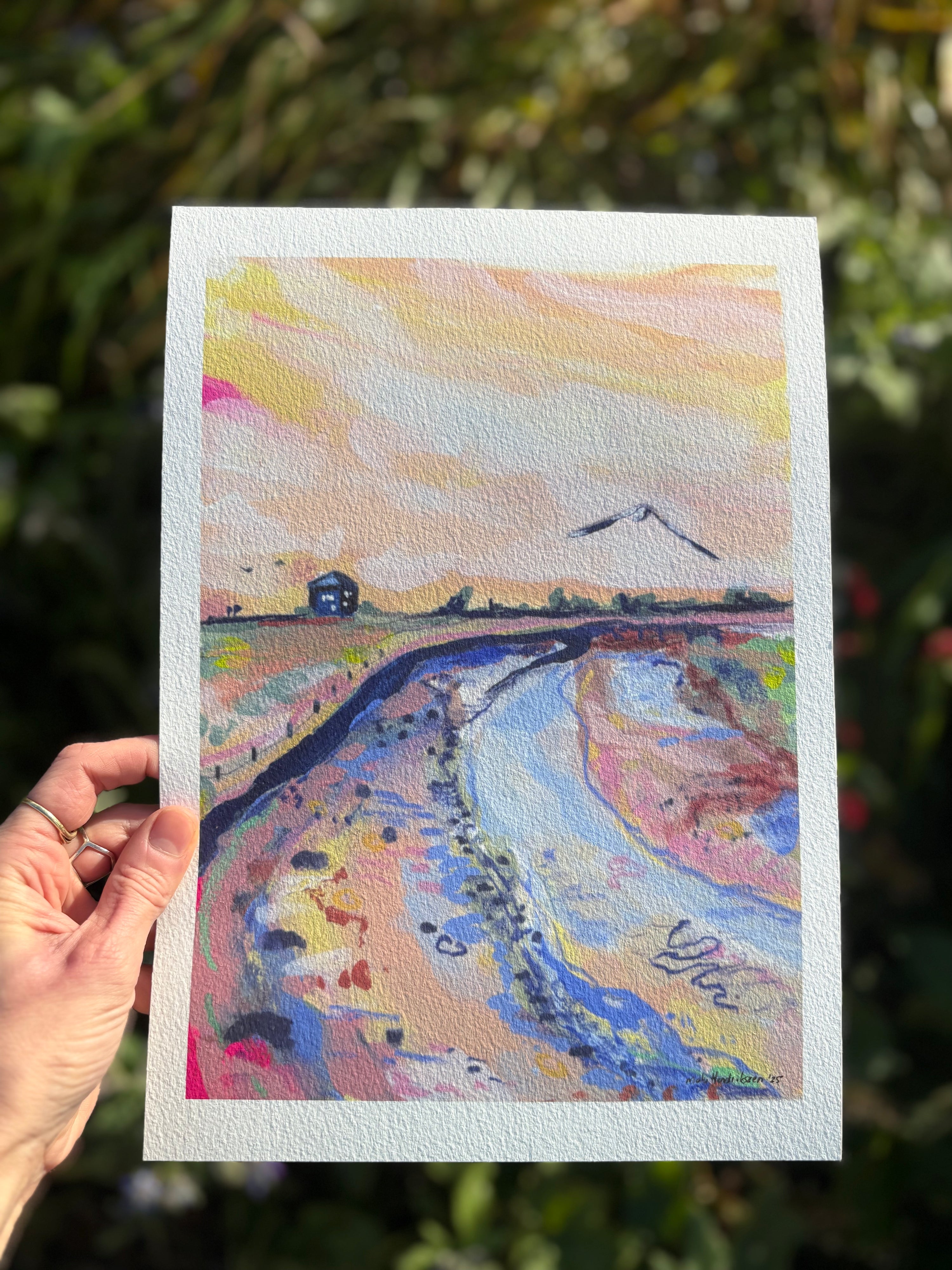 Hand holding a colorful landscape painting with a house and river.