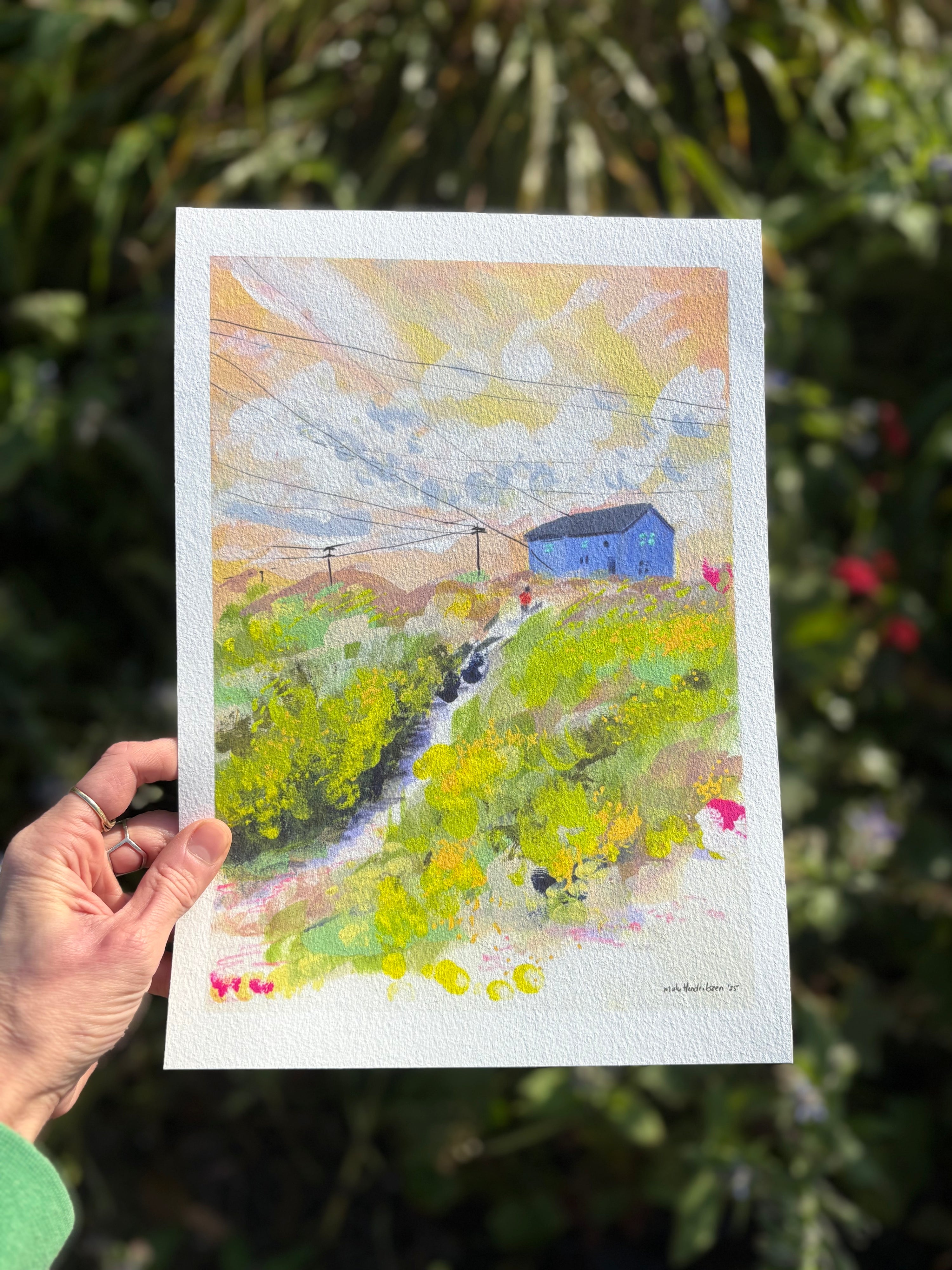 Hand holding a colorful landscape painting with a blue house against a green background