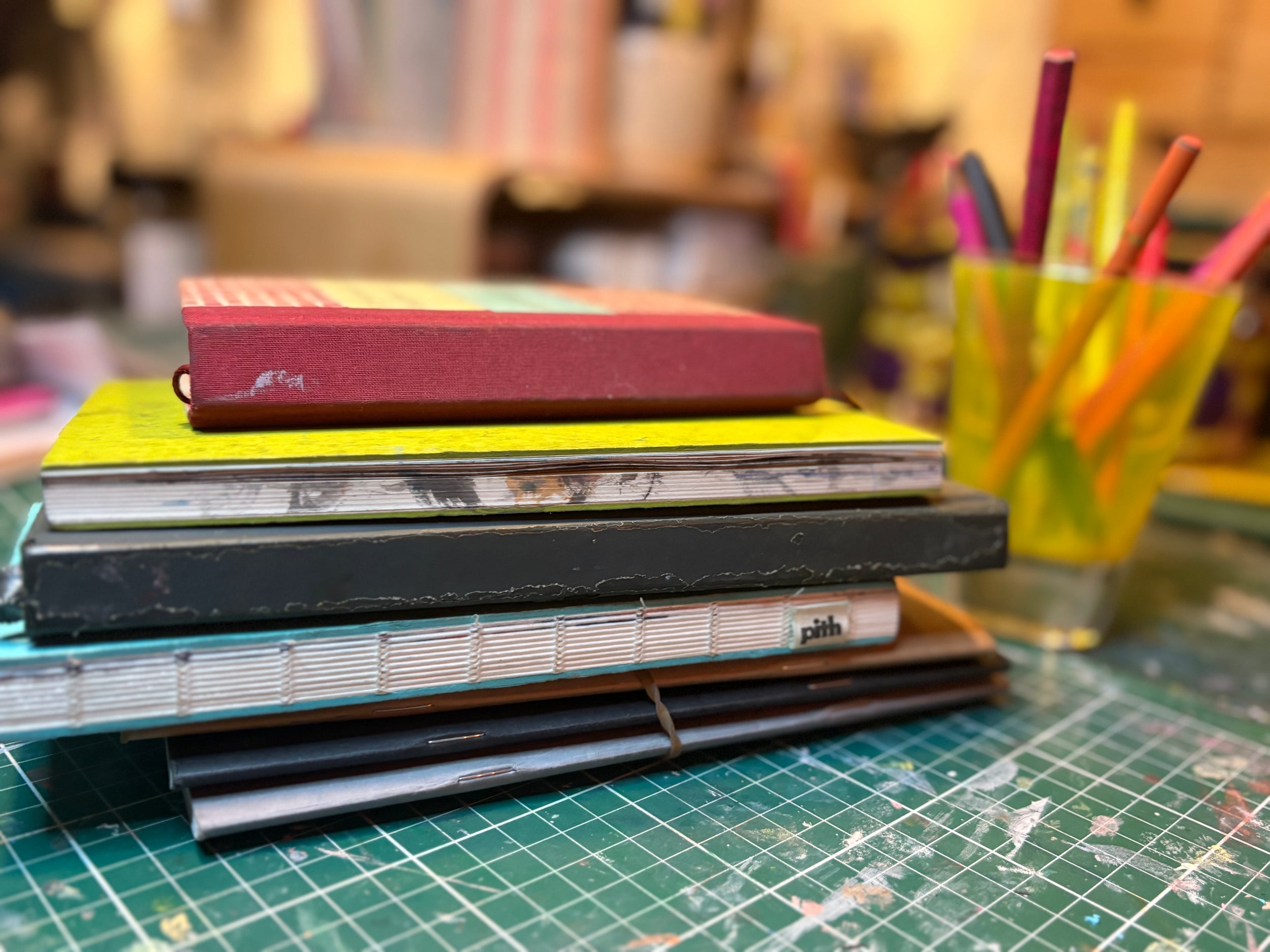 Stack of colorful sketchbooks on a green cutting mat with blurred stationery items in the background.