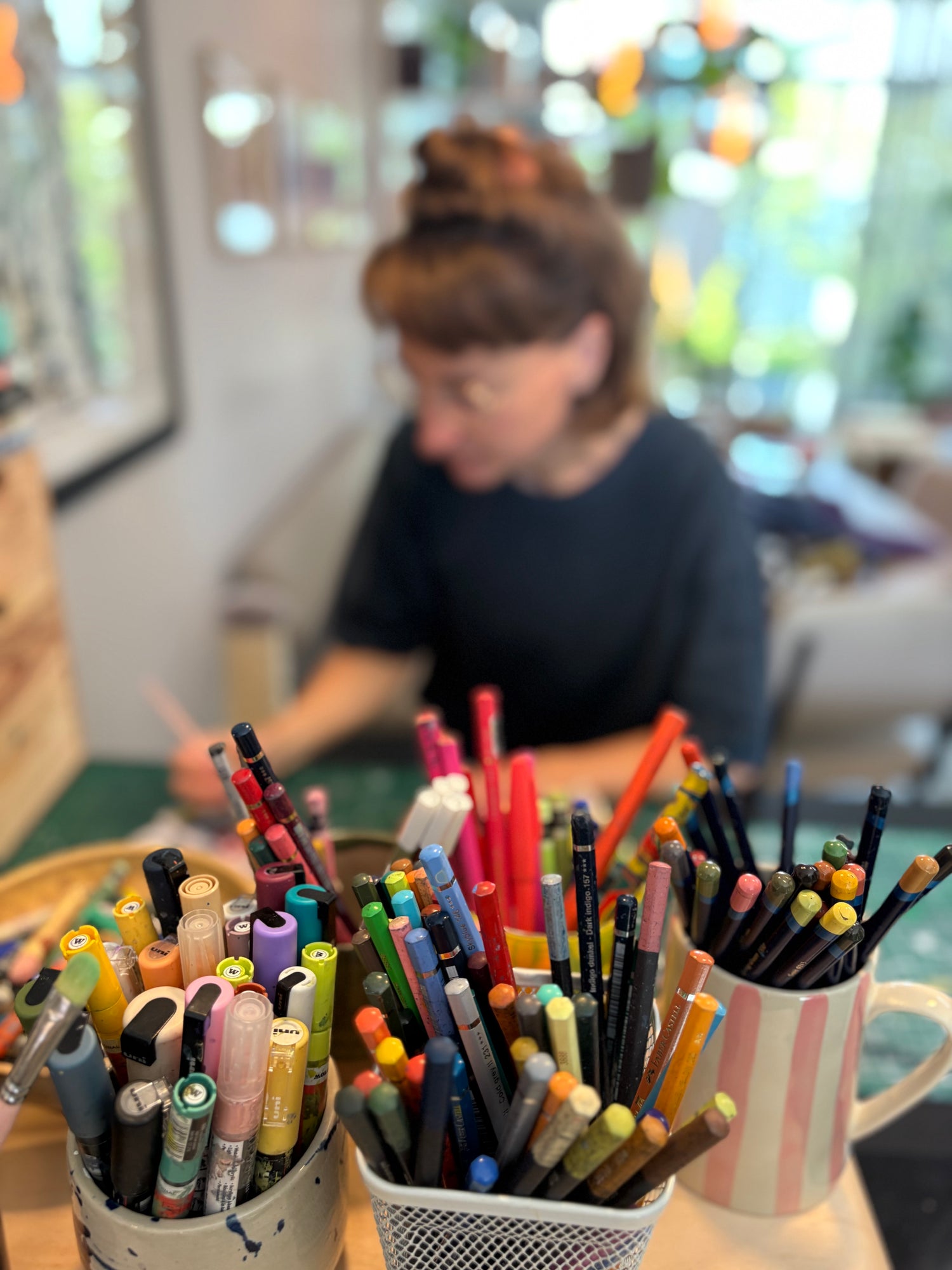 Colorful markers and pens in cups with Malu blurred  in the background