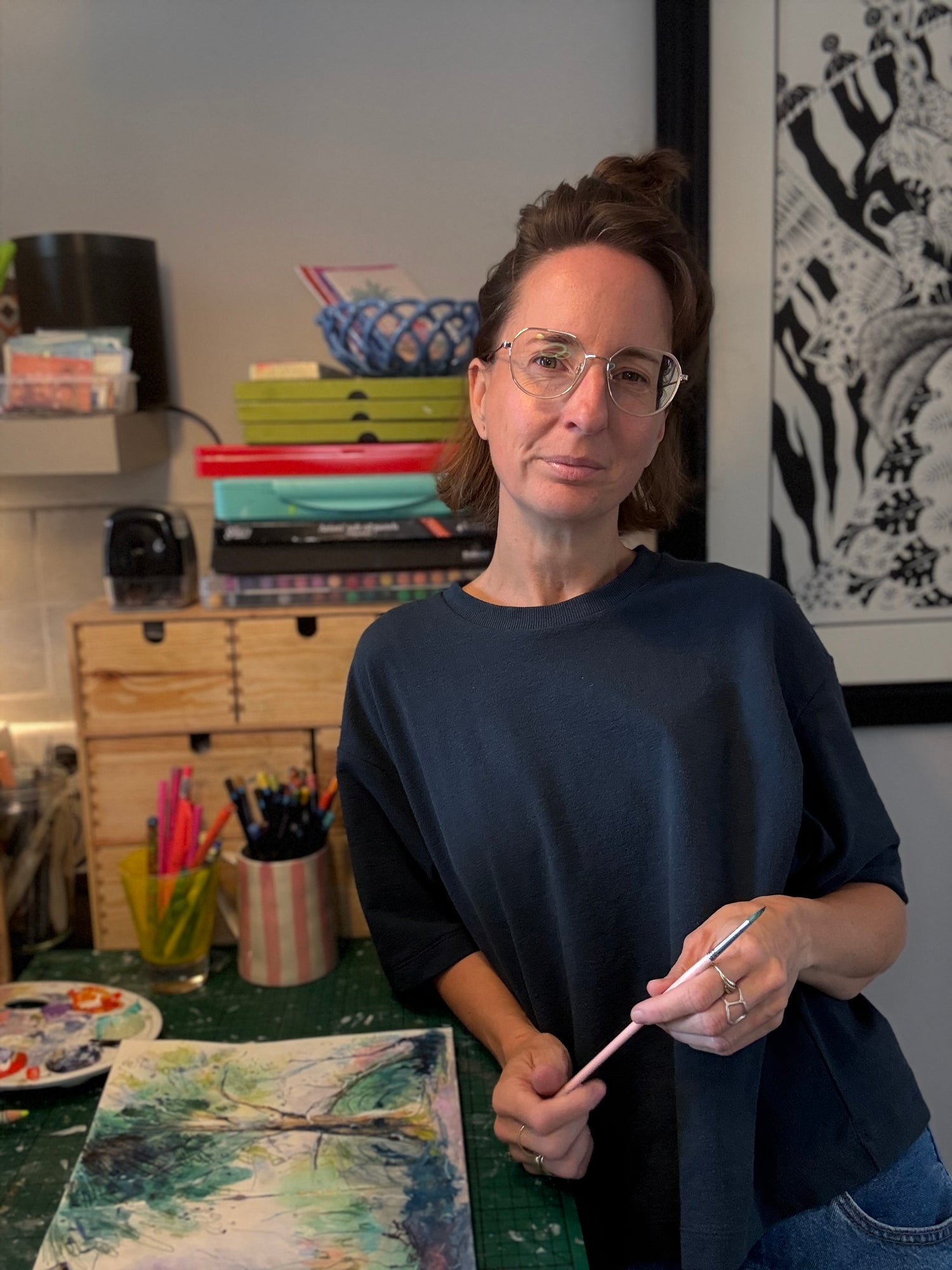 Malu holding a paintbrush in an art studio with art supplies and a painting on a table.