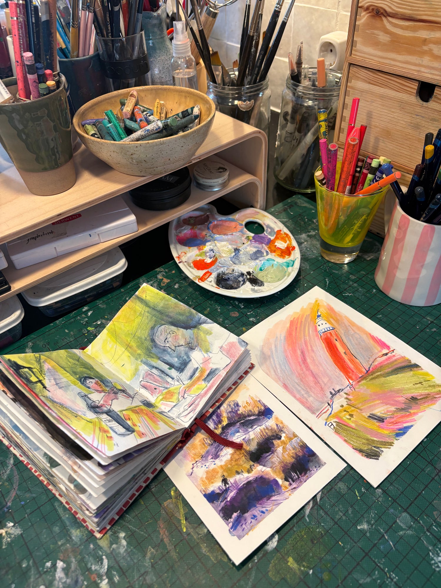 Artistic workspace with paintbrushes, paint palette, and watercolor paintings on a green surface.
